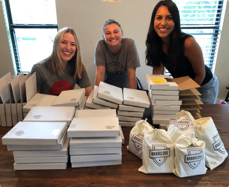 Three women smile while sitting at a table organized with boxed items and drawstring bags labeled "BraveLove.