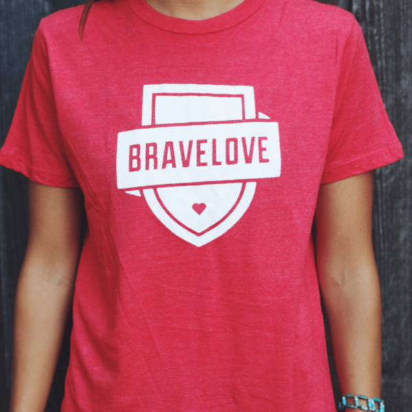 Person wearing a Red T-Shirt featuring a white shield design and the word "BRAVELOVE" printed on it, standing against a dark background.