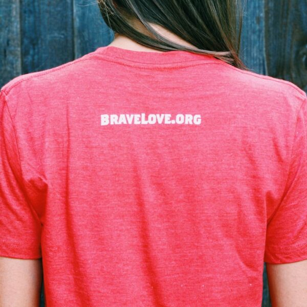 A person wearing a Red T-Shirt with "BRAVELOVE.ORG" printed on the back, standing in front of a wooden wall.