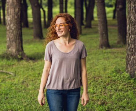 A woman with red hair and glasses stands in a forest clearing, wearing a gray t-shirt and jeans, smiling and looking to the side.