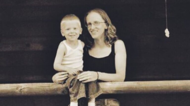 A woman with glasses and a young boy, both smiling, sit on a wooden railing in front of a dark background.