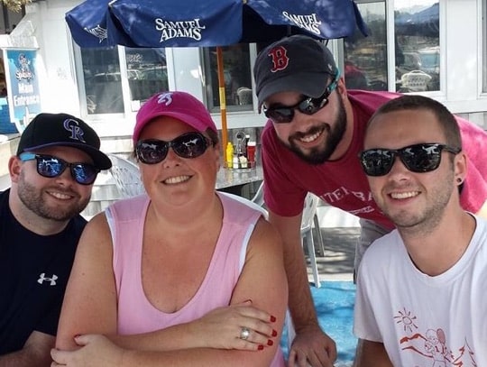 Group of four smiling individuals wearing casual attire, sunglasses, and hats, posing at an outdoor restaurant under umbrellas.