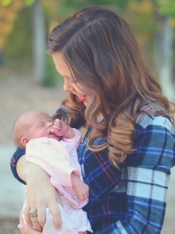 A woman with long brown hair, wearing a plaid shirt, lovingly holds a sleeping baby in her arms in a serene outdoor setting.