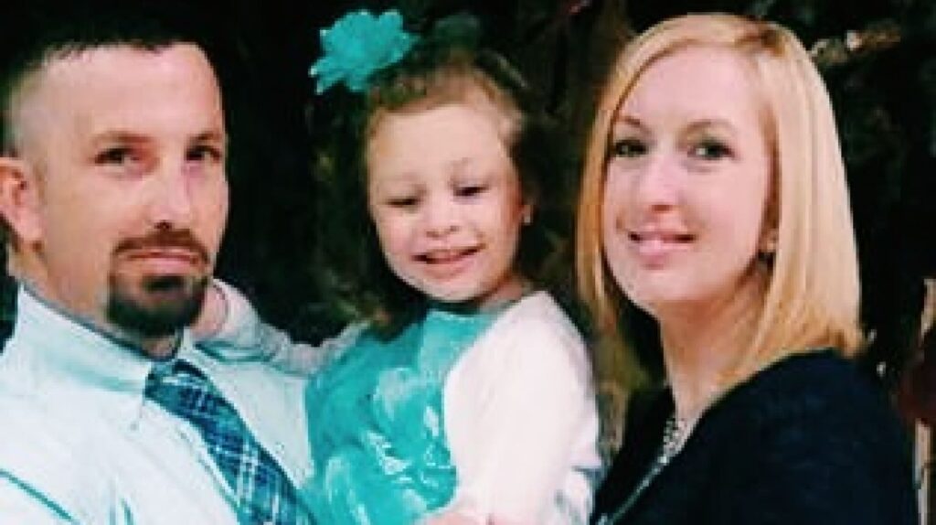 A man and woman smile while holding a young girl in a blue dress with a matching bow in her hair.