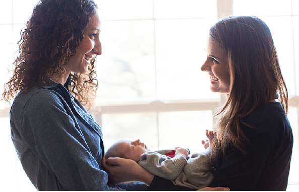 Two smiling women stand by a window, one holding a baby wrapped in a blanket.