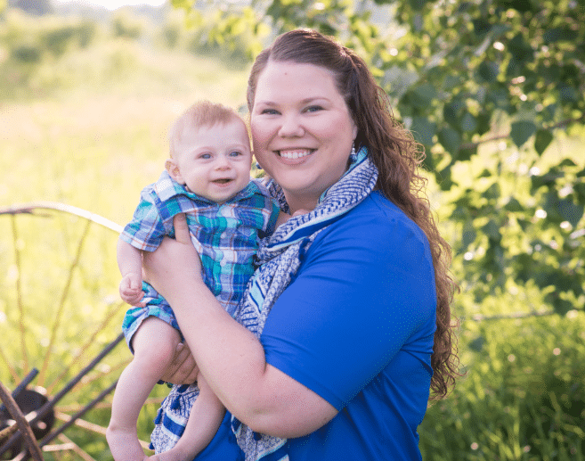 Woman in a blue dress smiling and holding a baby in a plaid outfit, standing outdoors with greenery and a rustic wagon wheel behind them.