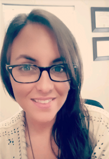 A woman with long dark hair wearing glasses is smiling at the camera. She is indoors, and framed documents are visible on the wall behind her.