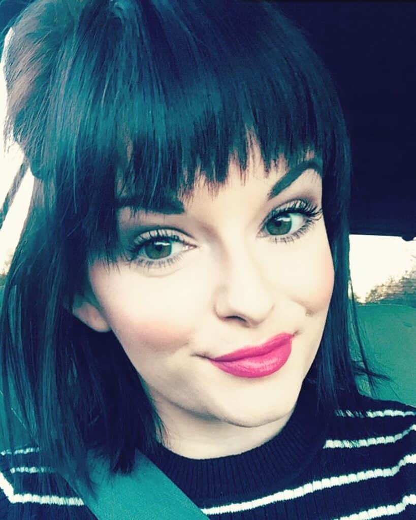 Woman with dark hair and bangs, wearing a striped top and pink lipstick, smiling at the camera while sitting in a car.