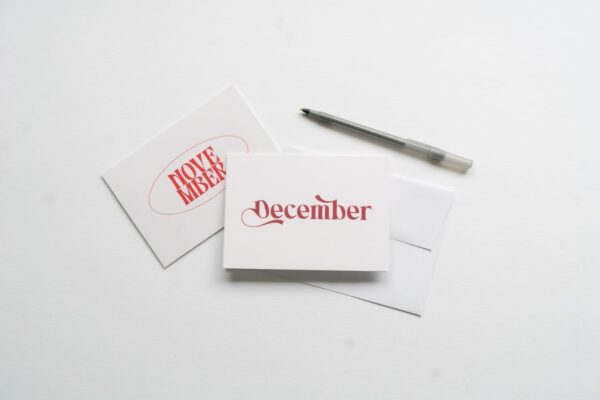 Two cards from the "New! Months of the Year Stationary Set" on a white surface, one displaying "November" and the other "December" in red font, accompanied by a pen.