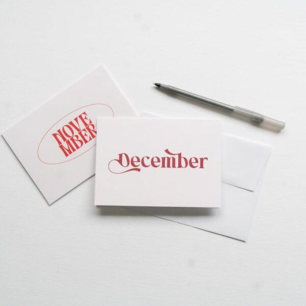 Two cards from the "New! Months of the Year Stationary Set" on a white surface, one displaying "November" and the other "December" in red font, accompanied by a pen.