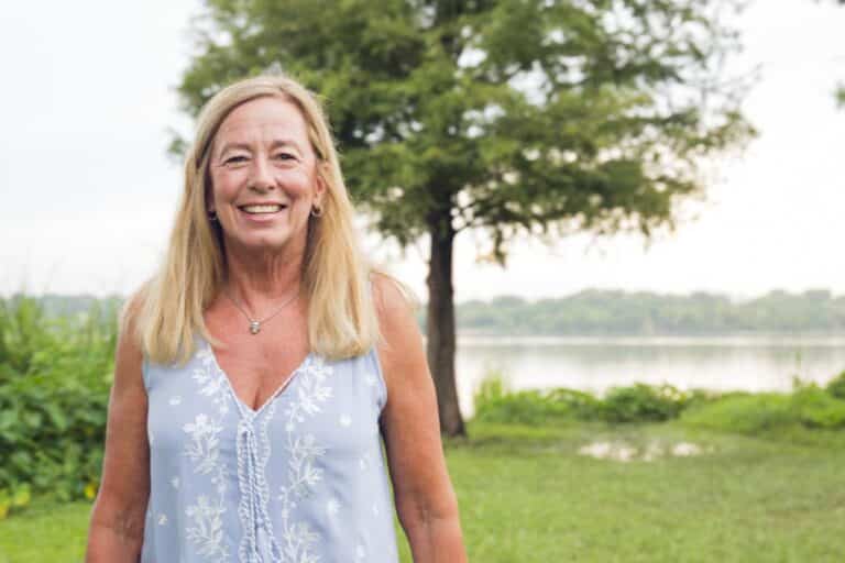 A smiling woman with long blonde hair stands outside in front of a tree and lake, wearing a light blue sleeveless top with white floral prints.