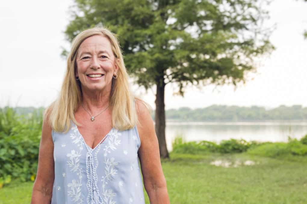 A smiling woman with long blonde hair stands outside in front of a tree and lake, wearing a light blue sleeveless top with white floral prints.