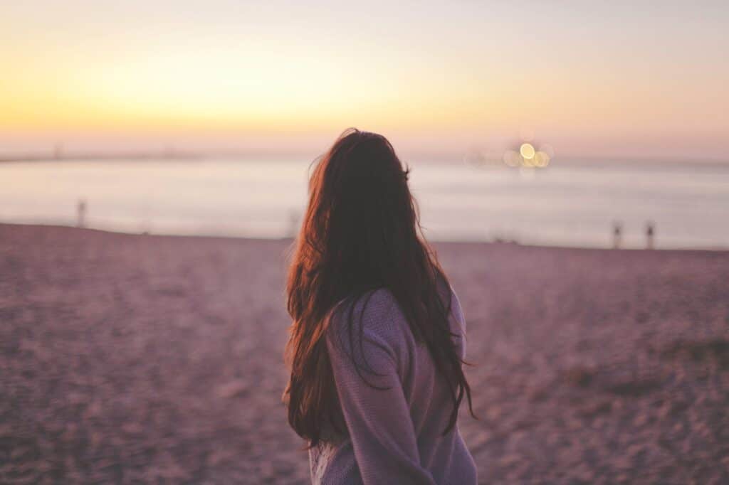 A person with long hair stands on a beach at sunset, looking out at the ocean.