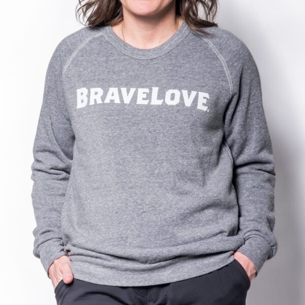 A person wearing a Sweatshirt with "BRAVELOVE" printed on the front, hands in their pockets, stands against a white background.