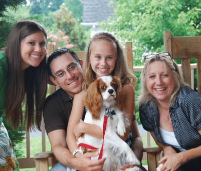 Four people and a dog sit on a porch, smiling at the camera. The child holds the dog, a Cavalier King Charles Spaniel, on her lap.