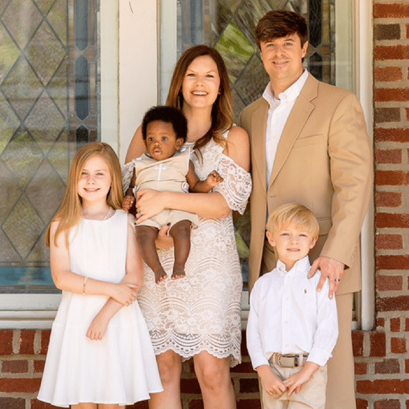 A family of five, dressed in white and beige, stands in front of a brick wall and window. The mother holds a baby while the other children stand nearby.