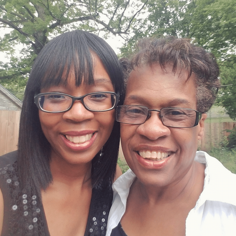 Two women wearing glasses, smiling, standing closely together outdoors with trees and a fence in the background.