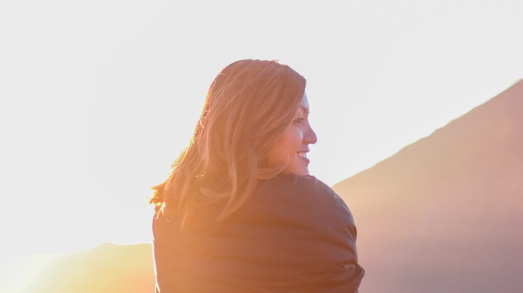 A person with shoulder-length hair smiles while looking at the sunrise or sunset, with a mountain in the background.