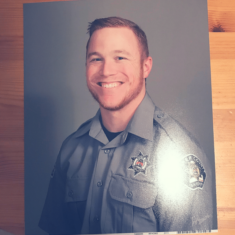 Smiling man with short light hair in a law enforcement uniform standing against a neutral background.