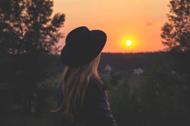 A person wearing a black hat looks at a colorful sunset surrounded by trees and distant houses.