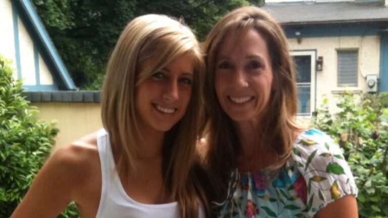 Two women smiling outdoors, one with long blonde hair in a white top, the other with long brown hair in a floral blouse.