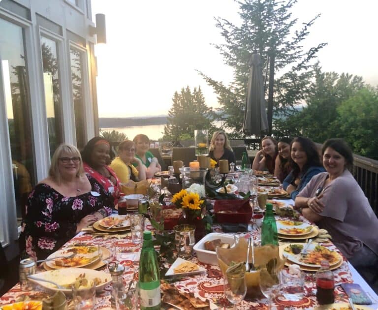 A group of women sitting around a long, decorated outdoor table, sharing a meal with a scenic view in the background.