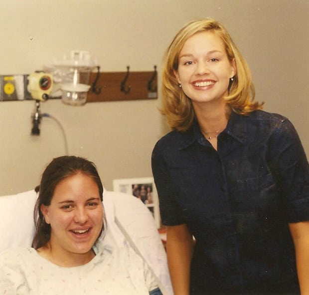 Two women smiling; one sitting on a hospital bed wearing a gown, the other standing beside her in casual attire with medical equipment nearby.