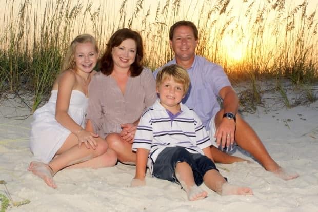 A family of four sits on a sandy beach at sunset, smiling. The mother and daughter wear dresses while the father and son wear shirts and shorts.