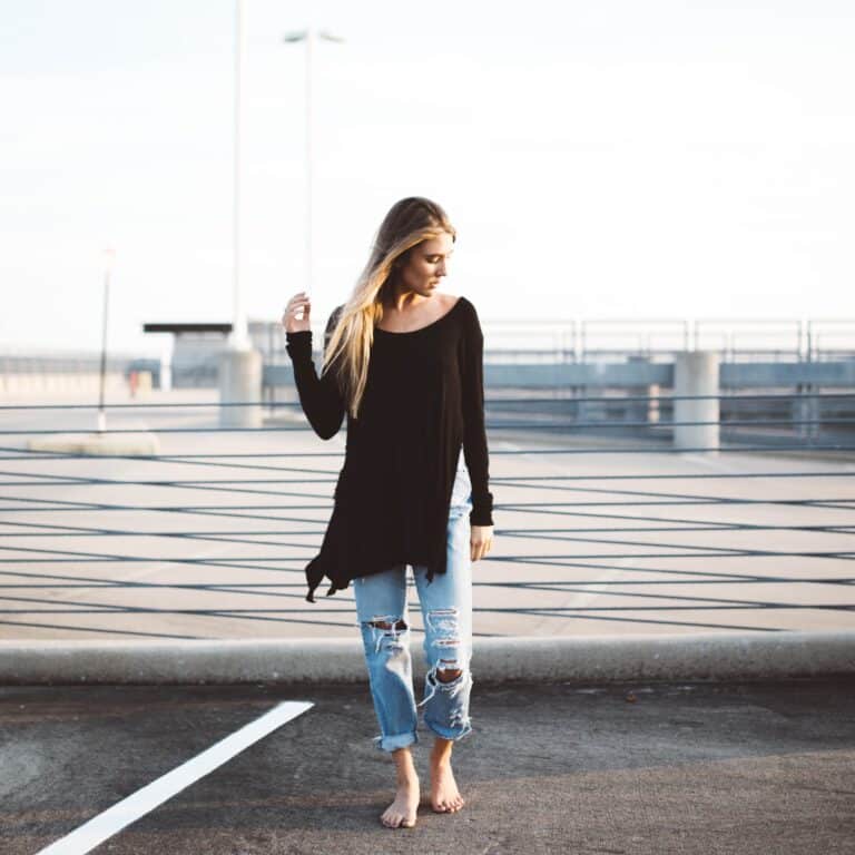 A woman with long hair stands barefoot on a rooftop wearing a loose black top and ripped jeans, looking down at the ground.