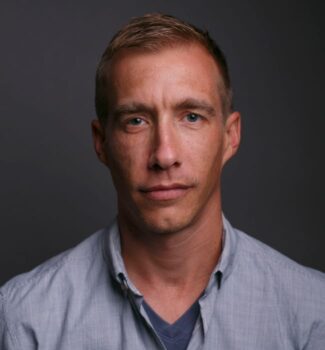 A man with short hair, wearing a light blue button-up shirt and a gray t-shirt, poses against a dark gray background.