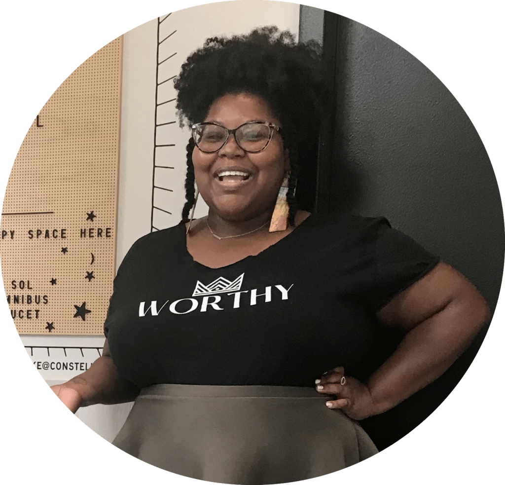 A smiling person with glasses and an afro wears a black "WORTHY" shirt while standing in front of a bulletin board.