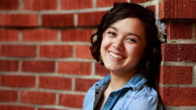 Young woman smiling while leaning against a brick wall, wearing a denim jacket and a white flower in her dark, curly hair.