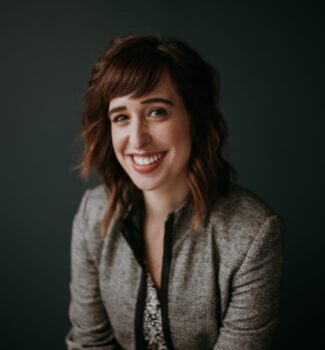 A woman with wavy brown hair smiles while wearing a gray blazer and patterned blouse against a dark background.