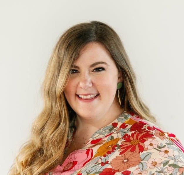 Woman with long, wavy hair smiles at the camera wearing a floral patterned top against a plain white background.