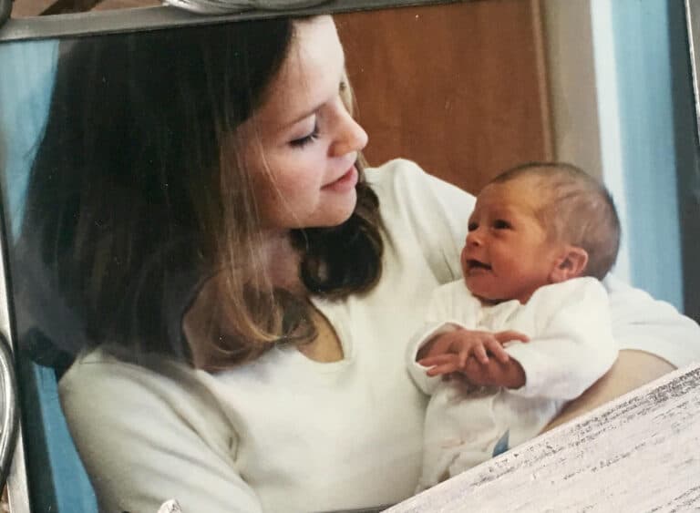 A woman holds a newborn baby in her arms, both gazing at each other lovingly inside a room.