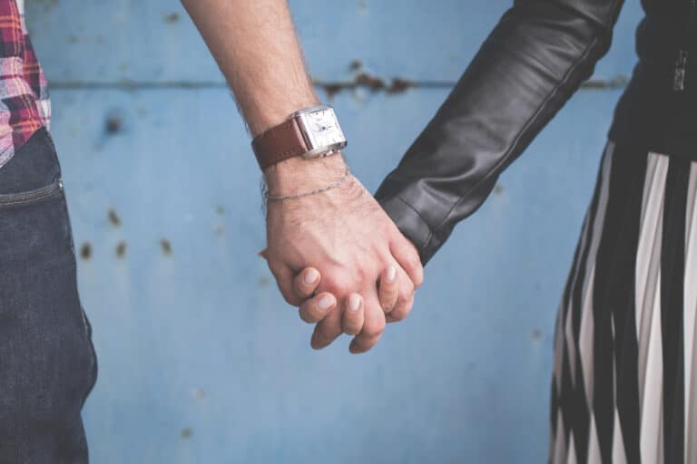 Two people holding hands against a blue background; one wears a leather jacket, the other a plaid shirt and a wristwatch.