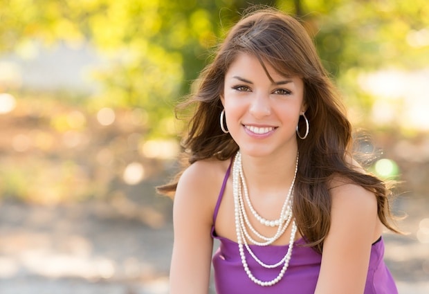 A woman with long brown hair, wearing a purple top and layered pearl necklace, smiles outdoors with greenery in the background.