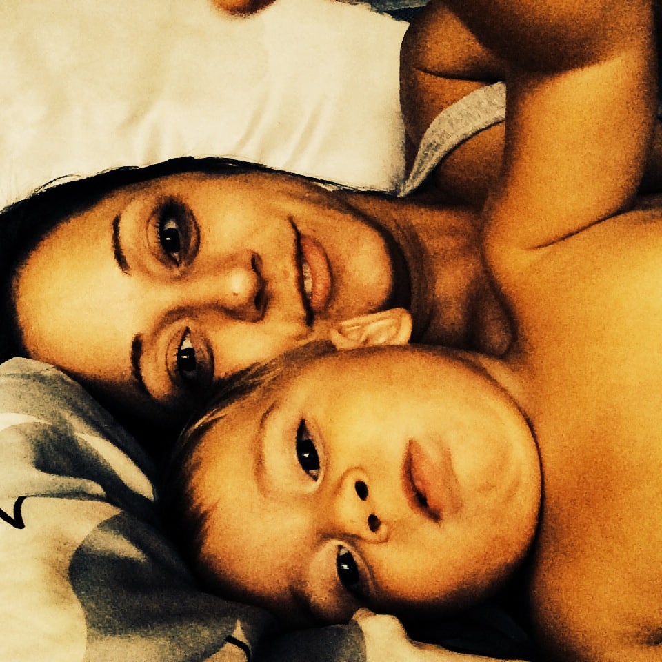 A woman and a young child lie closely together on a bed, looking at the camera. They appear relaxed and content.