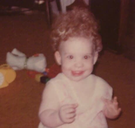 A young child with curly hair smiles and looks at the camera, seated on the floor with toys scattered in the background.