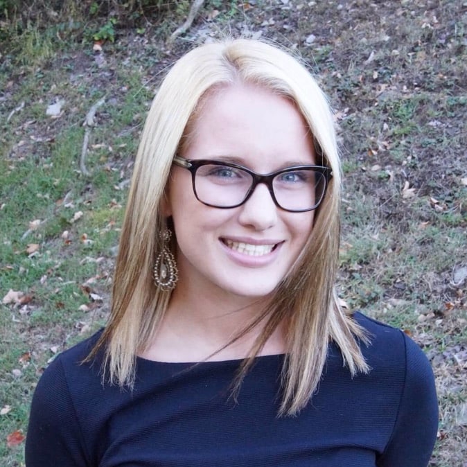 A person with long blonde hair, glasses, and dangling earrings wearing a black top, smiling outdoors on a grassy background.