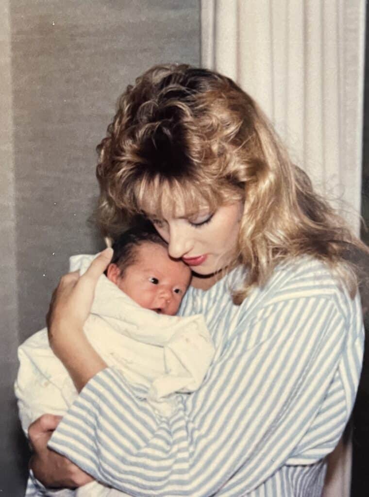 A woman with blonde hair wearing a striped shirt cradles a newborn baby wrapped in a blanket, both looking off to the side.