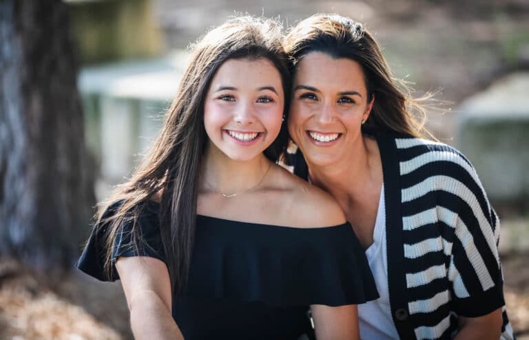 Two women smile brightly, posing outdoors. One wears an off-shoulder black top, and the other a striped cardigan over a white shirt.