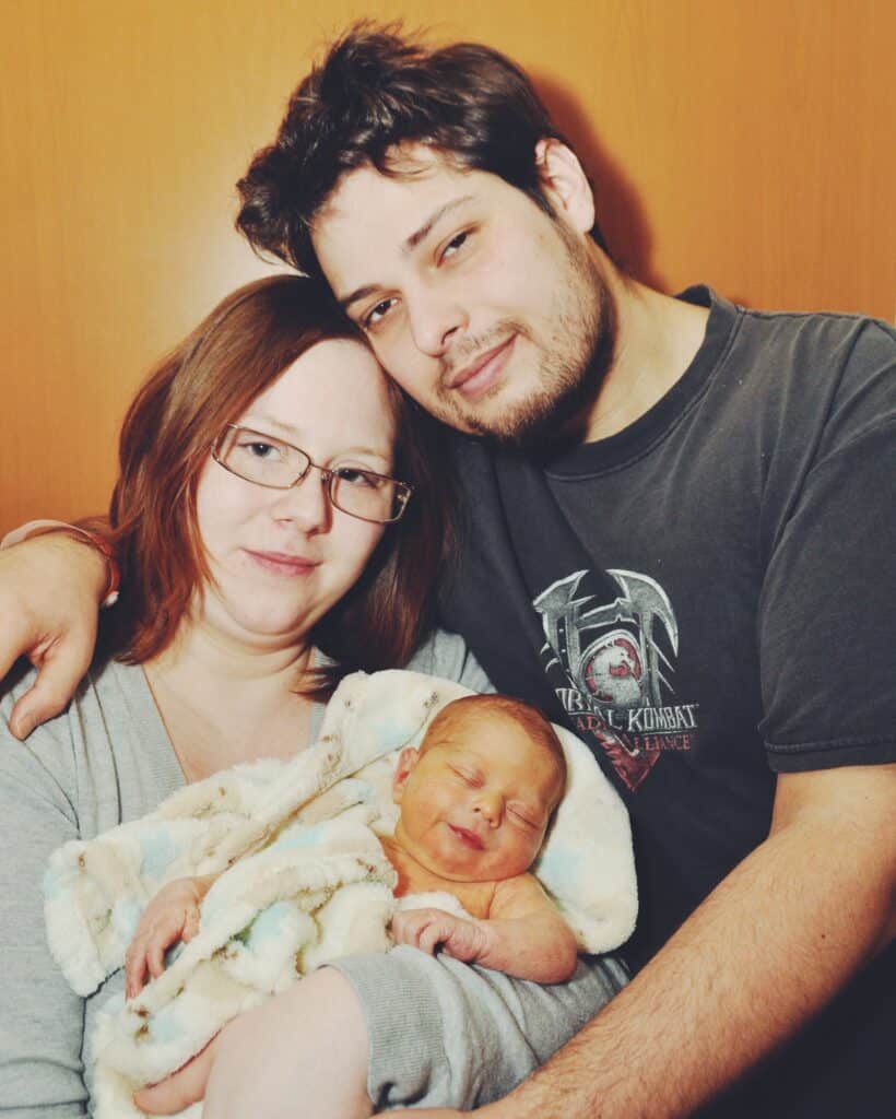 A couple smiling and holding a newborn baby wrapped in a blanket. The man has his arm around the woman. They are in front of a wood-like background.