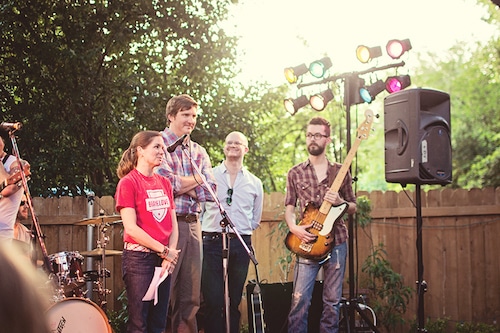 People standing on an outdoor stage with microphones and musical instruments, under colorful stage lights during a daytime event.