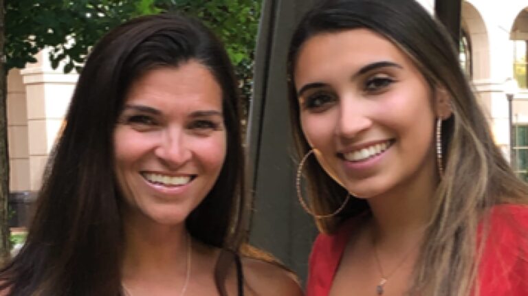 Two smiling women with long hair, one wearing a red top and hoop earrings, standing outdoors against a background of trees and buildings.