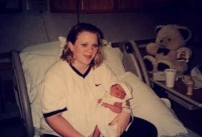 A woman sitting on a hospital bed holds a newborn baby wrapped in a white blanket, with a teddy bear and various items in the background.