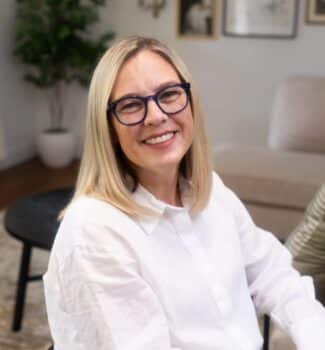 A smiling person with glasses and blonde hair, wearing a white shirt, sits in a cozy, well-decorated indoor space with framed pictures.