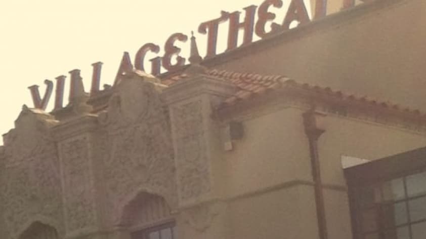 A building with the sign "VILLAGE THEATRE" on the rooftop, featuring intricate architectural details and a sunlit exterior.