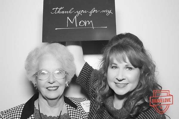 Two women smile; one holds a sign that reads "Thank you for my Mom." A "BRAVELOVE" logo is at the bottom right. The photo is in black and white.