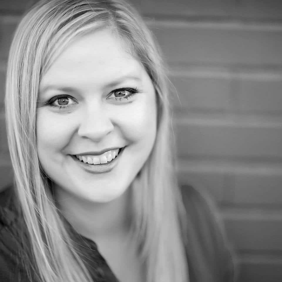 A woman with long blonde hair smiles, looking at the camera, against a brick wall background in a black and white image.